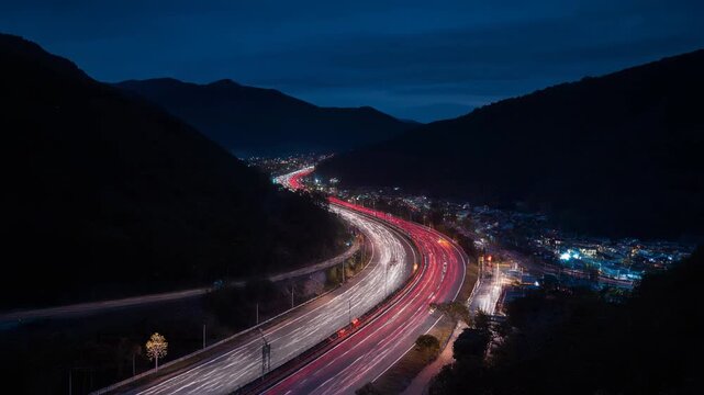 Carretera serpenteante con estelas de luz de veh&iacute;culos entre monta&ntilde;as oscuras al anochecer, con un pueblo iluminado en la distancia.