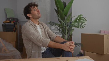 Handsome man sitting in new home surrounded by moving boxes and plants contemplating future decisions