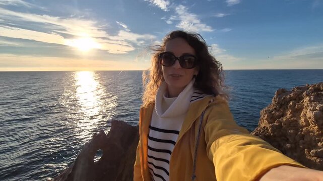 Woman travel sunset, talking on selfie camera from a stunning rocky cliff overlooking the ocean at golden hour.