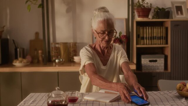 Medium shot of cheerful elderly Caucasian woman with gray hair, living with chronic diabetes ailment in old age, placing smartphone to sensor patch, checking blood glucose and writing in notebook