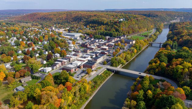 An aerial view of a small town nestled in a river valley, surrounded by dense, colorful autumn foliage and spanned by a bridge over the tranquil canal.