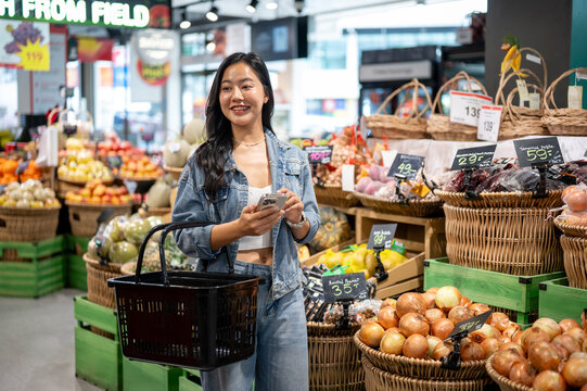 Asian woman holding shopping basket look away from phone walking in vegetable section at supermarket