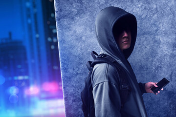 Hacker in hoodie with black bag hiding behind dark grunge wall on the street at night city