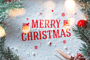Merry Christmas greeting written on snowy surface with pine branches, red ornaments and holiday lights. Winter festive background for greeting cards.