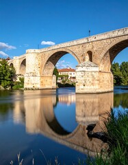 Fototapeta premium Stone arched bridge over river with reflections on water, under blue sky