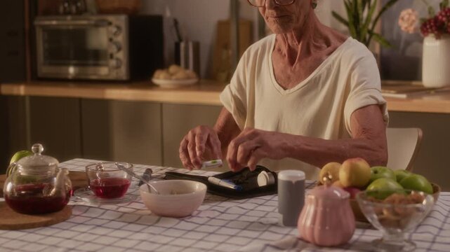 Medium shot of senior Caucasian woman with grey hair sitting at kitchen table after meal of porridge and fruit, preparing diabetes test kit to measure blood sugar, taking out meter and insulin pen