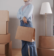 A young woman's body in a knitted sweater and jeans carries a jumbo paper shopping bag among a pile of cardboard boxes.
