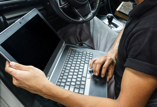 Unrecognizable auto mechanic using a laptop with a blank screen inside a car. Vehicle diagnostics, tuning, or modern auto repair service concept.