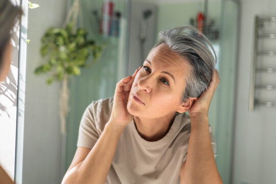 European white-skinned middle-aged woman looks anxiously in the bathroom mirror. Gray hair, wrinkles