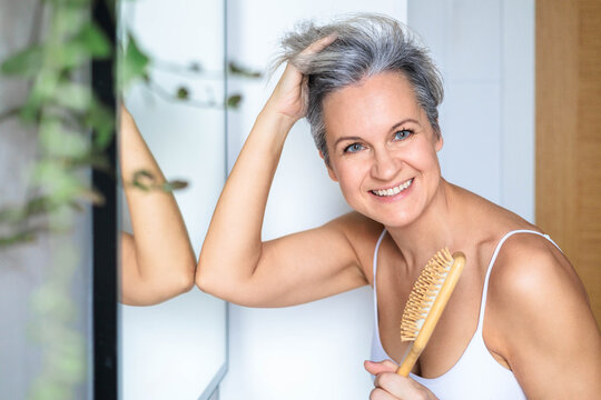 European beautiful and smiling middle-aged woman combing her gray hair near the mirror