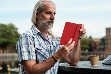Senior man enjoying a book and coffee by the canal
