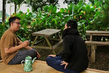 A father and his teenage son sitting together at an outdoor garden table, enjoying a quiet moment surrounded by lush greenery in a peaceful natural setting.