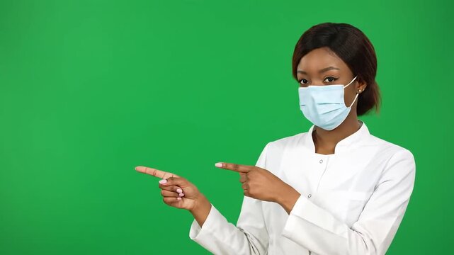 A young African American female doctor or nurse in a medical mask points to the side presenting information or a product on a green screen background ideal for healthcare and promotional content.