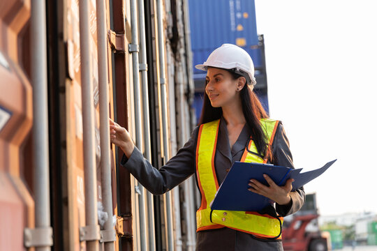 Female inspector in safety vest and helmet checking shipping container data for logistics and transportation industry, ensuring import-export accuracy and freight security. - Powered by Adobe