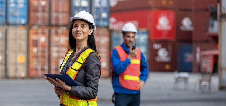 Confident female logistics supervisor in safety vest holding clipboard at shipping yard with male worker in background managing container cargo operations and communication.