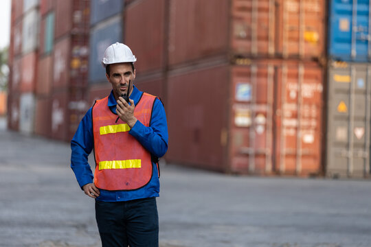 Male logistics worker in safety vest and helmet using walkie-talkie at container yard, coordinating shipping operations, communication, cargo management for freight industry.