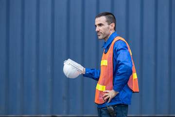 Side view of male logistics worker holding safety helmet and wearing reflective vest, standing near...