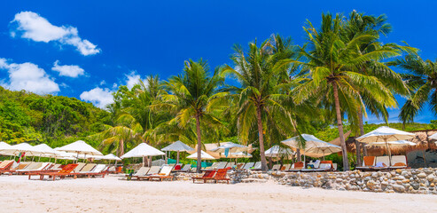 luxury tropical resort with sun loungers and umbrellas under palm trees on beach by sea in summer. Landscape panorama with a view of exotic ocean shore. A tourist destination for holidays