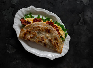 Delicious breakfast, tapas, snack - roti tortilla with omelet, bacon and green salad on a dark background, top view