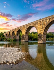 Fototapeta premium An arched, stone bridge spans a calm river at sunset. The sky is a gradient of colors, reflecting off the water. Lush foliage lines the banks