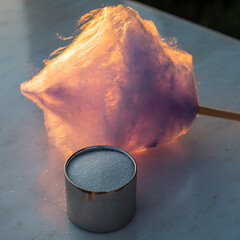 A fluffy pink and purple cotton candy illuminated by warm light sits on a marble surface next to a small silver container.