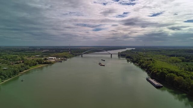 Aerial hyperlapse of the Pentele Bridge spanning the Danube River near Dunavecse, Hungary, with cargo ships navigating below under dramatic cloudy skies.