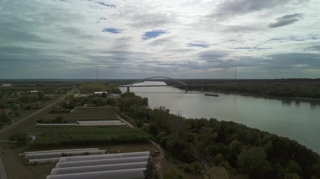 Drone ascending from Dunavecse, Hungary, revealing the Pentele Bridge spanning the Danube River under overcast skies with greenhouses and riverside vegetation in the foreground.