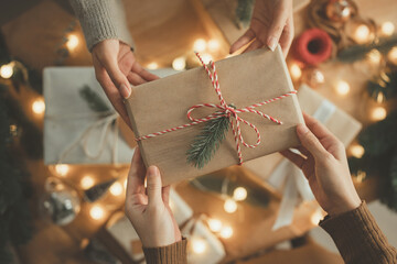 Hands exchanging Christmas gift box wrapped in brown paper with pine decoration and red white...