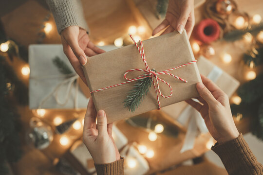 Hands exchanging Christmas gift box wrapped in brown paper with pine decoration and red white string, festive holiday season celebration with warm lights, friendship and happiness in home, New year - Powered by Adobe