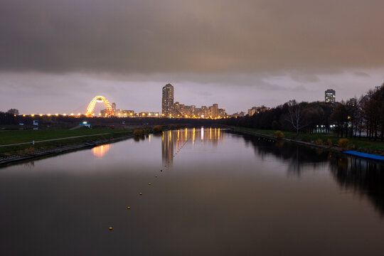 A dramatic twilight view of the Moscow skyline and the illuminated Zhivopisny Bridge, with calm reflections in the Krylatskoye rowing canal under a heavy, overcast sky.