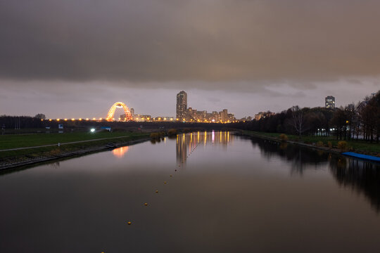 A dramatic twilight view of the Moscow skyline and the illuminated Zhivopisny Bridge, with calm reflections in the Krylatskoye rowing canal under a heavy, overcast sky.