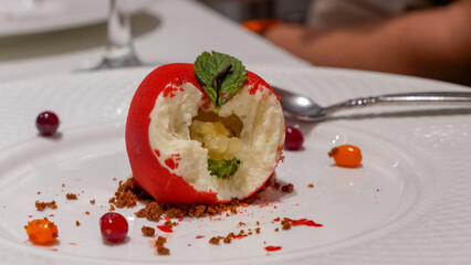 Red dessert shaped like an apple, featuring creamy filling and garnished with mint leaves, displayed on a white plate with colorful berries