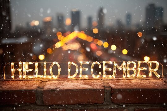 Hello December formed by glowing string lights on a brick wall. Blurred background of city lights at dusk, snowflakes falling.