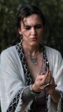 A close-up of a beautiful brunette holding a smoking palo santo stick during a magical ritual against a green forest backdrop. The woman, dressed in boho style, meditates and dances in the forest.