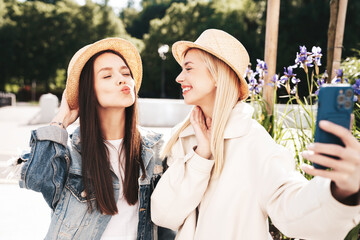 Two young beautiful smiling hipster female in trendy summer clothes. Carefree women posing in the street. Positive models having fun outdoors at sunny day. Cheerful and happy. In hat, take selfie