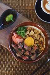 A close-up shot of a premium steak rice bowl topped with a raw egg yolk, crispy garlic chips, pickled ginger, and fresh herbs.