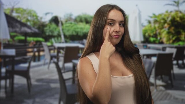 Woman finger to cheek on restaurant terrace street in sleeveless top revealing cleavage and long hair; subtle confidence.