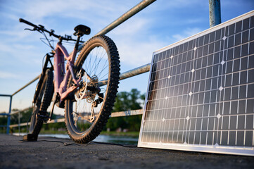 Focus on solar panel against backdrop of blue sky and distant trees. Pink electric mountain bike parked by lakeside railing, connected to solar panel for charging.