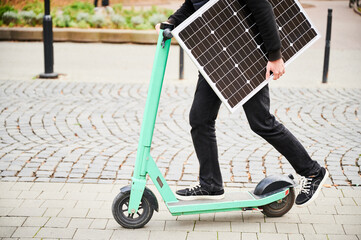 Cropped view of man rides electric scooter while carrying solar panel. Integration of solar power as sustainable energy source for charging electric scooters, promoting eco-friendly transportation.