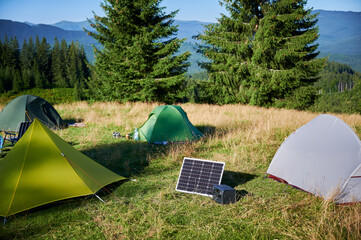 Tourist tent with solar panel and portable power station nearby in grassy field. Scene surrounded by rolling hills, tall pine trees under clear blue sky, showcasing eco-friendly camping setup.