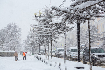 View of a snow-covered city street during a heavy snowfall. Lots of snow on the sidewalk. Janitor removes snow. Cars are stuck in a traffic jam. Low visibility during a snowstorm. Snowy winter weather