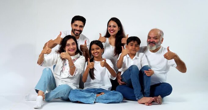 Indian family showing success thumbs up sitting isolated on seamless white background, cheerful grandparents, parents and two kids smiling together, expressing happiness, confidence, and achievement