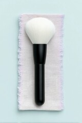 A fluffy white makeup brush with a black handle rests on a piece of pink fabric against a light blue background.