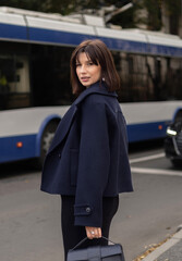 Fototapeta premium Pregnant woman poses on an urban street. Female in black dress and short coat walks by a trolleybus and white building, leans on massive columns, checking her bag. Stylish city look.