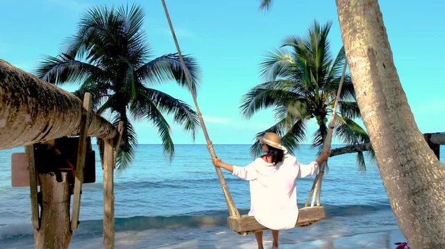 A woman enjoys a peaceful moment swinging on a beach swing, surrounded by palm trees and the tranquil ocean waves of Koh Kood Island, Thailand. Perfect for showcasing tropical paradise vibes.