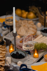 Autumn picnic in the park with a table full of food, pancakes, ham, cheese, and tea. Someone pours sparkling wine into a glass. Cozy setup with candles, fairy lights, pillows, and blankets.