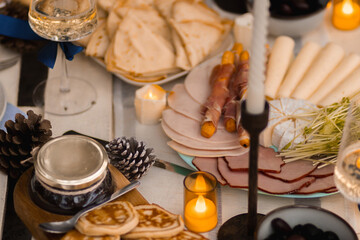 Autumn picnic in the park with a table full of food, pancakes, ham, cheese, and tea. Someone pours sparkling wine into a glass. Cozy setup with candles, fairy lights, pillows, and blankets.