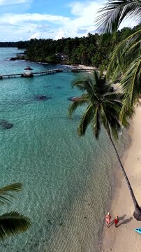 Two people enjoy a peaceful stroll along the sandy beach of Koh Kood Island, Thailand, surrounded by crystal clear waters and lush palm trees under a bright blue sky.