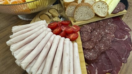 High-quality close-up of a delicious charcuterie board featuring assorted cured meats, sliced turkey/ham rolls, salami, smoked beef, cheese slices, cherry tomatoes, and fresh bread.