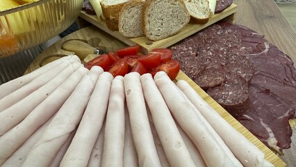 High-quality close-up of a delicious charcuterie board featuring assorted cured meats, sliced turkey/ham rolls, salami, smoked beef, cheese slices, cherry tomatoes, and fresh bread.